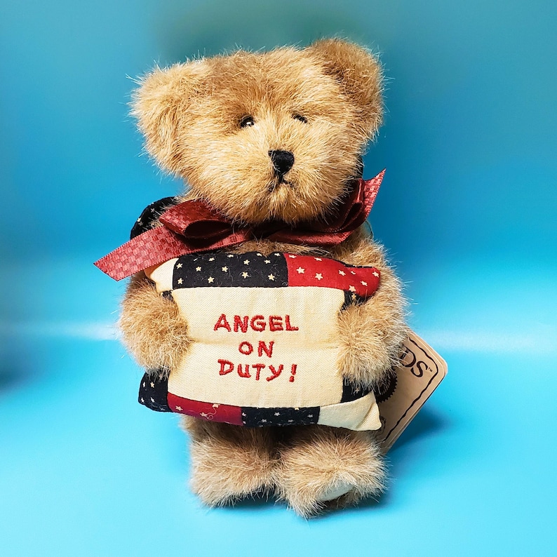 Peut inclure: Un ours en peluche brun avec un n&oelig;ud bordeaux et un coussin brod&eacute; avec "ANGEL ON DUTY!". Le coussin pr&eacute;sente un motif patchwork avec des motifs d'&eacute;toiles rouges, noires et blanches. Une petite &eacute;tiquette est attach&eacute;e.