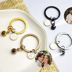 Può includere: Portachiavi in oro, oro rosa, argento e nero, ciascuno con un anello rotondo e un ciondolo. I ciondoli includono una piccola foto e un disco bianco con una lettera. I portachiavi sono esposti su un piatto bianco con sfondo di giornale.
