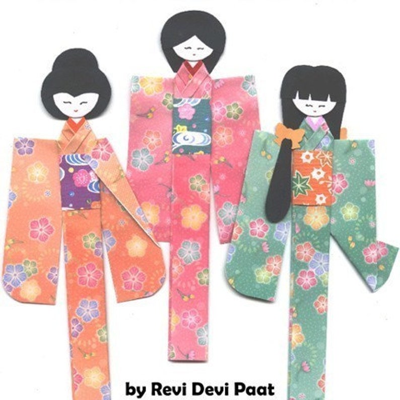 May include: Three paper dolls dressed in colorful kimonos with floral patterns. The dolls have black hair and simple facial features. The text "by Revi Devi Paat" is at the bottom of the image.