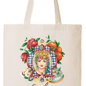 May include: Cream-colored tote bag featuring a watercolor illustration. The design includes a woman's portrait, surrounded by pomegranates, oranges, and pastries. The word "Sicilia" is written in a script font below the image.