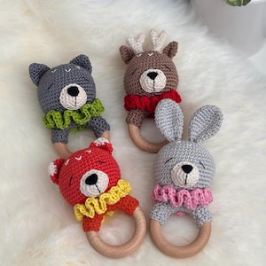 May include: Four handmade crochet baby rattles in the shape of animals: a bear, a deer, a rabbit, and another bear. Each has a wooden ring and a ruffled collar. The bear is red, the deer is brown, the rabbit is gray, and the other bear is gray.