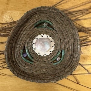May include: Handmade woven basket with a central seashell design. The basket is brown with green accents and a white scalloped edge around the shell. Decorative elements include small purple beads.