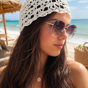 Puede incluir: Un gorro de ganchillo blanco con un intrincado patrón floral calado. La prenda está confeccionada con hilo fino y presenta un delicado diseño de borde festoneado que se ajusta a la cabeza, mostrando una textura detallada y repetitiva similar al encaje.