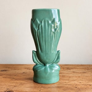 May include: A tall, cylindrical ceramic vase with a glossy, muted seafoam green glaze. The exterior features a raised, molded relief pattern of vertical, petal-like shapes that extend from the base upward toward the wide, open circular rim.