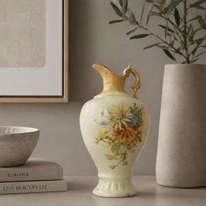 May include: A cream-colored ceramic pitcher featuring a glossy finish, a curved handle, and a decorative floral illustration on the front. The design includes yellow, orange, and blue flowers with green leaves painted on the bulbous body of the vessel.