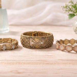 May include: Three gold-toned bracelets are displayed on a light wood surface. The bracelets vary in design, including a stretch bracelet with clear stones, a wide cuff with a geometric pattern, and a hexagonal link bracelet with pink accents.