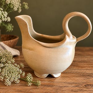May include: A cream-colored ceramic pitcher with a rounded body, a wide pouring spout, and a high, curved handle. The vessel features a smooth, glazed finish and rests on a small, scalloped base, showcasing a simple, functional design.