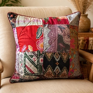 May include: A square throw pillow featuring a patchwork design of various batik fabrics. The pillow displays a mix of red, black, green, and purple panels with intricate floral and geometric patterns, finished with a dark red piped edge.