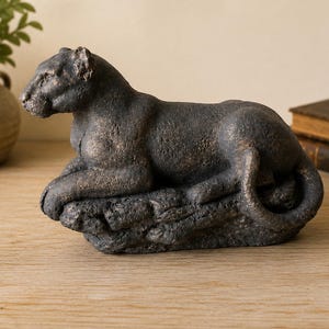 May include: A dark bronze-toned sculpture of a panther in a resting position, perched atop a textured, rocky base. The feline figure features a weathered, matte finish with subtle highlights, capturing the animal's muscular form and long tail.