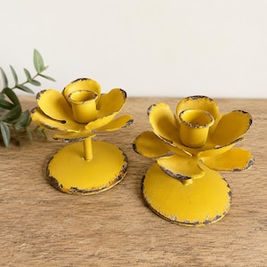 May include: A pair of yellow metal candle holders shaped like open flowers. Each holder features a circular base, a central stem, and layered petals with distressed, dark-colored edges around the top cup for a candle to be placed inside.