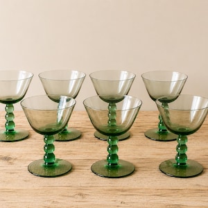 May include: Set of seven green-tinted glass coupe glasses. Each glass features a round base, a stem with a series of spherical accents, and a wide, shallow bowl. The glasses are arranged on a light-colored wooden surface.