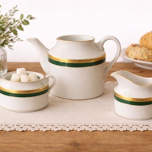 May include: A white porcelain tea set with a teapot, creamer, and sugar bowl. Each piece has a green and gold band. A plate of baked goods and a vase of flowers are in the background. The set is on a white lace doily.