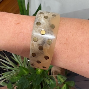 May include: A wide, beige and brown marbled bracelet with brass-colored circular studs. The bracelet is worn on a person's arm, with green plants in the background. The bracelet is a statement piece.