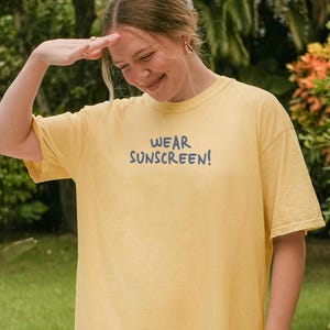 May include: A yellow t-shirt with the text "WEAR SUNSCREEN!" in blue. The shirt is worn by a person outdoors, with a hand raised to shield the sun. The background includes green plants and a stone patio.