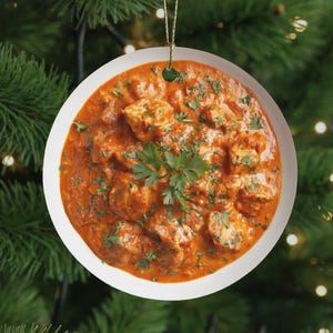 May include: A round, white Christmas ornament featuring a vibrant image of chicken tikka masala. The dish is a rich orange color, garnished with fresh green herbs, and presented in a white bowl. A gold string is attached for hanging.