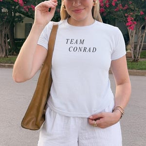 May include: A woman wearing a beige baseball cap with a crest, a white t-shirt with "TEAM CONRAD" printed in black, and white shorts. She is also wearing gold hoop earrings and a brown shoulder bag.