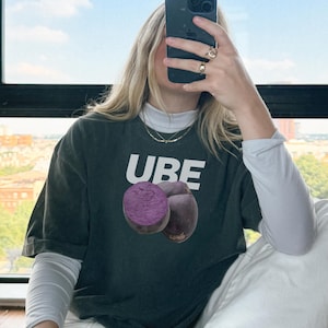 May include: A person wearing a dark grey t-shirt with a white graphic of two purple ube fruit and the word "UBE" printed in white. The person is wearing white pants and white sneakers.