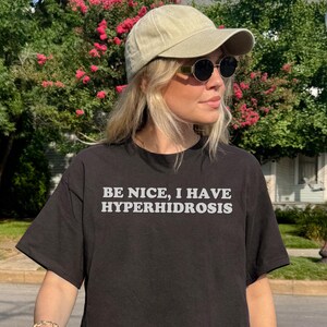 以下が含まれることがあります： 黒いTシャツを着て、白い文字で「BE NICE, I HAVE HYPERHIDROSIS」と書かれた人。ベージュのベースボールキャップとサングラスをかけています。その人は家の前の歩道に立っています。