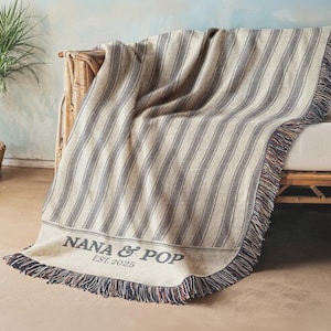 Puede incluir: Una manta a rayas beige y gris sobre un sofá. La manta tiene un borde de flecos con acentos rojos, blancos y azules. El texto "NANA & POP EST. 2025" está impreso en la manta.