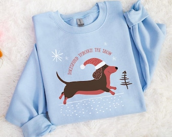 Peut inclure: Un sweat-shirt bleu clair avec un motif de teckel festif portant un bonnet de Père Noël et courant dans la neige. Le texte dit "Dachshund Through The Snow". L'image comprend des décorations lumineuses de Noël colorées.