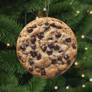 Op de afbeelding: Een ronde kerstversiering die lijkt op een chocolate chip cookie. Het ornament is lichtbruin met donkerbruine chocoladeschilfers. Het hangt aan een gouden draad tegen een achtergrond van een groene kerstboom.