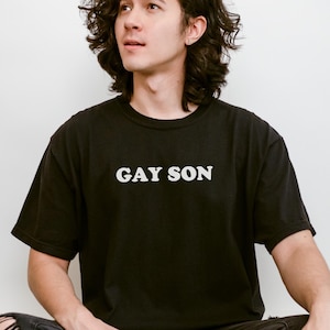 May include: A person wearing a black t-shirt with the text "GAY SON" in white letters. They are sitting with their legs crossed and looking up.