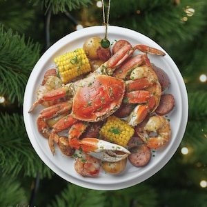 Könnte beinhalten: Weißer Keramik-Weihnachtsschmuck mit einem Meeresfrüchte-Boil. Der Schmuck zeigt eine Krabbe, Maiskolben, Kartoffeln, Würstchen und Garnelen auf einem weißen Teller, der an einem goldenen Faden hängt.
