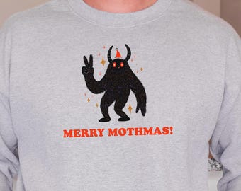 May include: A light grey sweatshirt featuring a black cartoon mothman character giving a peace sign and wearing a Christmas tree hat. The text "MERRY MOTHMAS!" is printed in red. A white mug with a red rim and a Christmas tree on a red truck is held.
