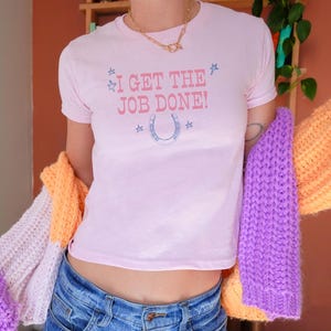 Puede incluir: Una camiseta rosa corta con el texto "I get the job done!" y un gráfico de herradura. La camiseta la lleva puesta una persona con un cárdigan de punto morado y naranja.