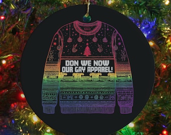 May include: A black Christmas ornament with a rainbow-colored sweater design. The sweater has the text "DON WE NOW OUR GAY APPAREL!" and festive details. The ornament hangs from a gold string against a backdrop of a Christmas tree.