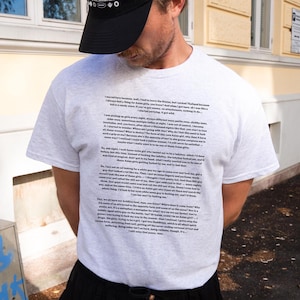 May include: A gray t-shirt with black text printed on the front. The text is a long paragraph about a person's experiences in Thailand. The person is wearing a black baseball cap.