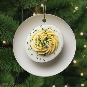 May include: A white ceramic ornament shaped like a deviled egg, with a swirl of yellow filling and green chives. The ornament hangs from a gold string against a backdrop of a green Christmas tree with twinkling lights. A unique holiday decoration.