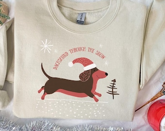 Peut inclure: Sweat-shirt beige avec un dessin d'un teckel portant un bonnet de Père Noël, courant dans la neige. Le texte "Dachshund Through The Snow" est au-dessus du chien. Un sapin de Noël et des flocons de neige sont également présents.