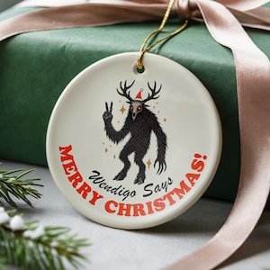 May include: A round white ceramic ornament featuring a black wendigo illustration wearing a small red Santa hat, making a peace sign. The text reads "Wendigo Says MERRY CHRISTMAS!" in red and black lettering. It hangs by a thin gold cord.