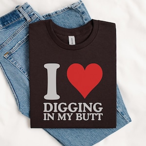 May include: A dark brown t-shirt with the phrase "I ❤️ DIGGING IN MY BUTT" in white lettering and a red heart graphic. The shirt is folded on top of a pair of blue denim jeans, all resting on a white surface.