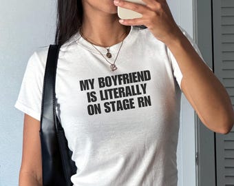 May include: White short-sleeved t-shirt with the text "MY BOYFRIEND IS LITERALLY ON STAGE RN" in black. The shirt is worn with a black shoulder bag and a patterned skirt. The image is taken indoors.