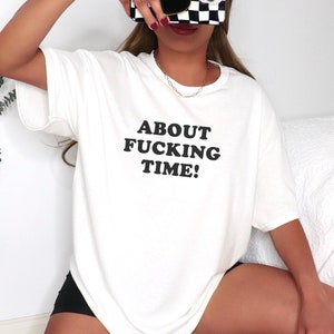 Może przedstawiać: Biała, oversizowa koszulka z czarnym nadrukiem "ABOUT FUCKING TIME!"