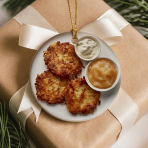 Könnte beinhalten: Ein runder weißer Keramikschmuck mit einer realistischen Darstellung von drei goldbraunen Kartoffelpuffern auf einem Teller, ergänzt durch zwei kleine Schalen mit weißer saurer Sahne und hellbraunem Apfelmus, an einer goldenen Schnur.