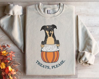 Puede incluir: Una sudadera de color beige claro con un gráfico de un perro negro y marrón asomándose de una calabaza llena de huesos blancos. El texto "Treats, Please." está impreso debajo de la calabaza.
