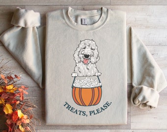 Puede incluir: Sudadera beige con una ilustración de un perro doodle blanco sosteniendo un cubo con forma de calabaza lleno de golosinas. El texto "TREATS, PLEASE." está impreso debajo. Las mangas están enrolladas.