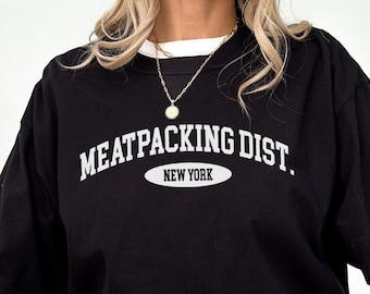 Puede incluir: Una camiseta negra con texto blanco que dice "MEATPACKING DIST. NEW YORK" en una fuente de estilo universitario.