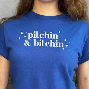 May include: A royal blue t-shirt with white text that reads "pitchin' & bitchin'" and white stars.