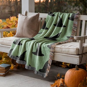 May include: A light green woven throw blanket featuring a repeating pattern of black witch hats. The blanket has a textured surface with fringed edges and is draped over a white wooden porch swing alongside a solid tan throw pillow.