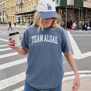 May include: A slate blue short-sleeved t-shirt with the text TEAM ALGAE printed in white block letters across the chest, paired with a white and slate blue baseball cap featuring the letters LA in dark blue embroidery and a white paper cup.