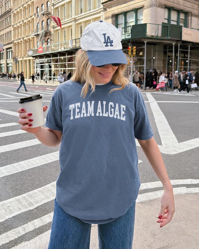May include: A slate blue short-sleeved t-shirt with the text TEAM ALGAE printed in white block letters across the chest, paired with a white and slate blue baseball cap featuring the letters LA in dark blue embroidery and a white paper cup.