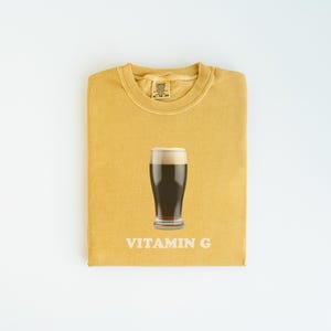 Pu&ograve; includere: Una maglietta gialla con un bicchiere di birra scura e la scritta "VITAMIN G" stampata su di essa.