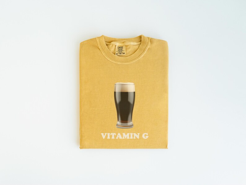 Pu&ograve; includere: Una maglietta gialla con un bicchiere di birra scura e la scritta "VITAMIN G" stampata su di essa.