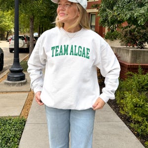 May include: A white crewneck sweatshirt featuring the text "TEAM ALGAE" in green capital letters across the chest, paired with a light-colored baseball cap and light blue denim jeans. The sweatshirt has long sleeves and a ribbed collar and cuffs.