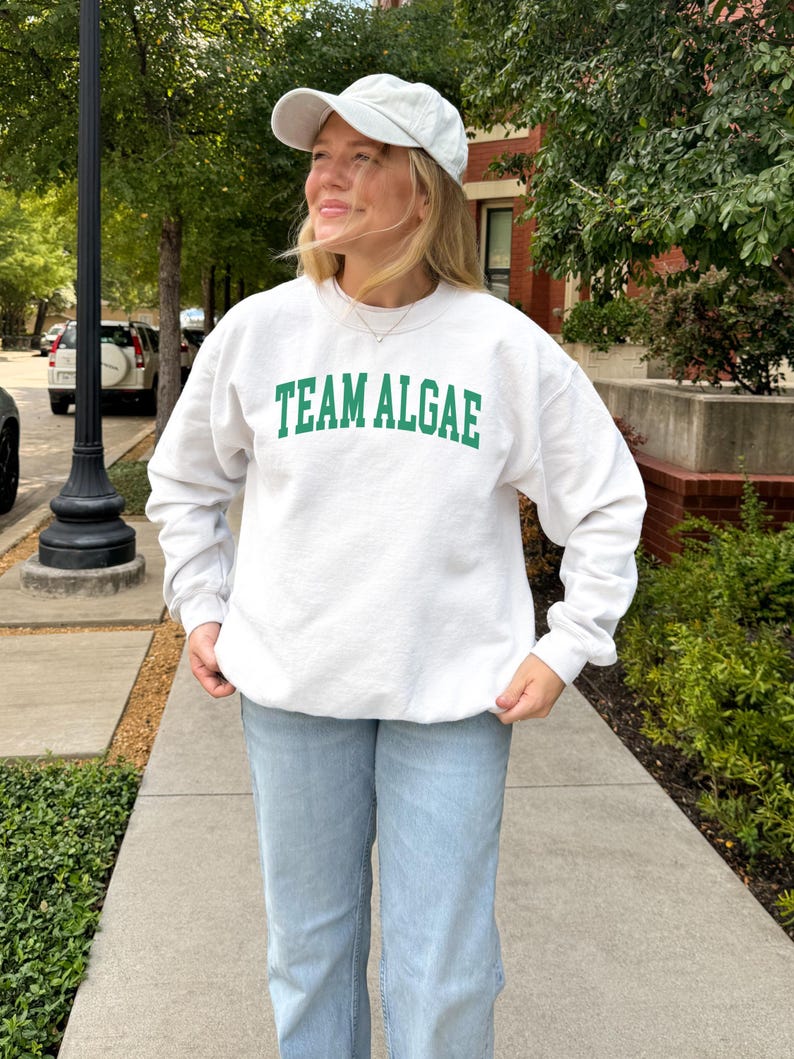 May include: A white crewneck sweatshirt featuring the text "TEAM ALGAE" in green capital letters across the chest, paired with a light-colored baseball cap and light blue denim jeans. The sweatshirt has long sleeves and a ribbed collar and cuffs.