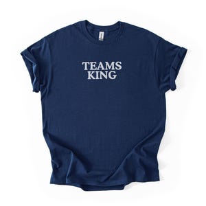 Könnte beinhalten: Marineblaues T-Shirt mit dem weißen Text "TEAMS KING". Das T-Shirt hat einen Rundhalsausschnitt und die Ärmel sind hochgekrempelt. Das T-Shirt besteht aus einem weichen Material.